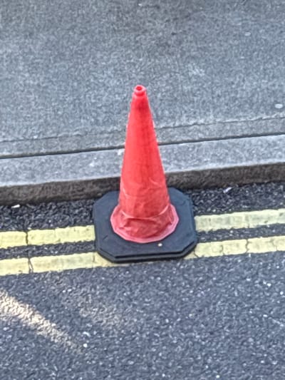 A single, bright red traffic cone stands on a grey asphalt road, positioned just beside a raised concrete curb. The cone's base is black and octagonal, and it is slightly wrinkled, suggesting it might be made of a flexible material. Two parallel yellow lines are painted on the asphalt in front of the cone, indicating a road marking. The background consists of more grey asphalt and the textured surface of the concrete curb. The scene appears to be an outdoor urban environment, likely a street. Without further context or discernible landmarks, it is impossible to pinpoint a specific location within the city of Cork, Ireland. There are no people, actions, or interactions visible in the image.
