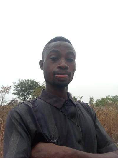 A young man with dark skin, short dark hair, and a light beard is featured in a selfie-style portrait. He is making a playful expression, sticking his tongue out. He wears a dark grey or black short-sleeved collared shirt with subtle vertical stripes and buttons down the front. An embroidered logo, possibly reading "UHM HOOD," is visible on the upper left arm/chest area of his shirt. A thin black chain necklace with a dark, rectangular pendant hangs around his neck. His arms are crossed in front of him.

The scene is outdoors, set against an overcast, light grey sky that occupies the upper portion of the frame. In the background, there are sparse trees with light green foliage and dry, light brown vegetation, suggesting a rural or undeveloped area. The lighting indicates it is daytime, but without direct sunlight. The location is consistent with an outdoor setting in Otokiti, Nigeria.