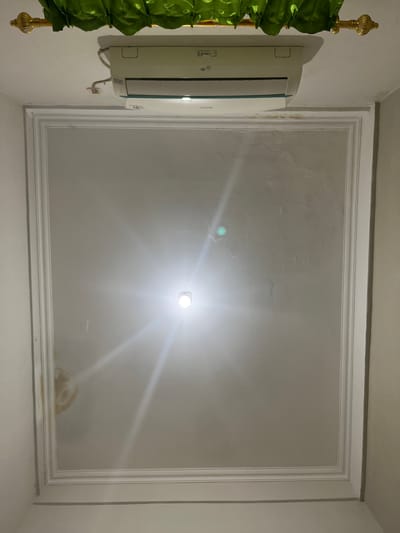 The image displays a ceiling with an air conditioning unit mounted on it. The ceiling has decorative molding, creating a recessed central area. A bright light fixture is visible in the center of the recessed area, causing glare and lens flares that obscure some details of the ceiling's surface, though some discoloration is present. Green drapery is visible at the top of the image, attached to a gold-colored rod. No people are present, and there are no indications of the specific location within Bandungrejo, Indonesia.