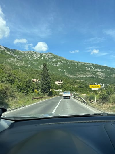The image depicts a view from inside a vehicle traveling on a road near Đurmani, Montenegro. A yellow sign for "Haj-Nehaj" is visible on the right side of the road. A mountain covered in vegetation rises on the left. Two vehicles are ahead on the road. The sky is blue with some clouds.