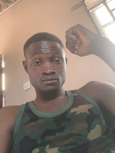 A dark-skinned male subject, with short dark hair and a neutral expression, is depicted in a close-up portrait, looking directly into the camera. He wears a green and brown camouflage-patterned sleeveless shirt with dark green trim. His right arm is raised with the hand clenched into a fist, positioned above his right shoulder. A silver-colored ring is visible on his right ring finger. The scene is indoors, located in Kado, Nigeria, against a light peach-colored wall. A white rectangular electrical fixture is visible on the wall to the subject's left. A window frame is partially visible in the upper right background, admitting bright light into the room.