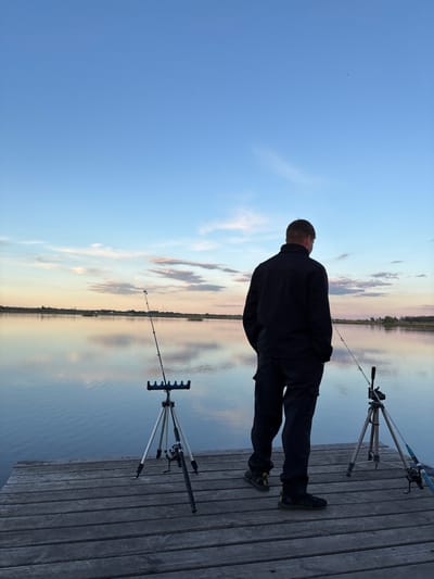 A man stands on a wooden pier, overlooking a calm body of water. Two fishing rods are set up on tripods on the pier beside him. He is wearing dark clothing and faces the water. The sky is a mix of blue and light, reflecting on the water's surface. This is likely at the city of Khryashchevka, Russia.
