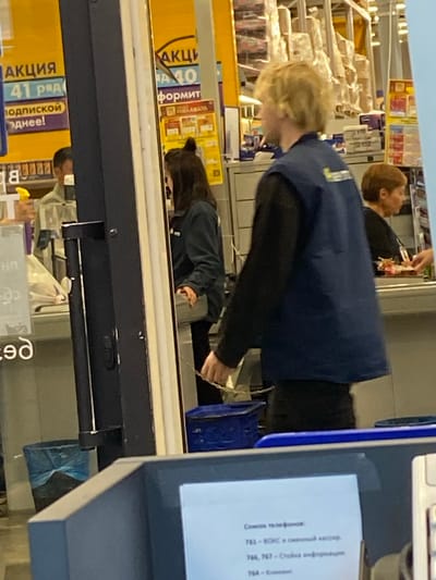 The media file captures an indoor scene, likely a retail environment such as a supermarket or department store, given the shelving filled with merchandise visible in the background. The primary subjects are two individuals, a young person with blond hair wearing a dark blue vest over a black long-sleeved shirt, and another person with dark hair wearing a dark long-sleeved shirt. Both individuals appear to be employees, possibly working at a checkout counter or stocking shelves.
The blond-haired individual is seen from the side, with their head bent down, possibly engaged in a task or looking at something on a lower level. Their vest has a visible logo or emblem on the shoulder. The dark-haired individual is also partially visible, their head down, suggesting they are also occupied with a task.
In the foreground, a blue plastic container is visible, and to the right, a portion of a counter with a screen displaying text is present. The text on the screen appears to be in Russian and lists telephone numbers and descriptions, likely related to customer service or store information. Some visible text on signs in the background includes "АКЦИЯ" (Promotion) and "40," "41," and "ПОДПИСКОЙ" (Subscription). These indicate promotional offers or information within the store.
The lighting suggests it's daytime, and the setting is indoors, offering no clues about the weather. The overall mood appears neutral, with individuals focused on their tasks. The location is identified as Arkhangelsk, Russia.