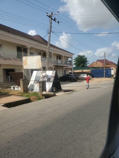 This image depicts a street scene in Kason, Nigeria. The foreground features an asphalt road with a concrete drainage channel on the left. In the mid-ground, a building with white and beige facade and balconies is visible. In front of the building, there is a display of ceramic tiles supported by brick pillars. A person in a red shirt and blue jeans walks away from the camera on the right side of the road. Behind the building, there are parked vehicles, including a black SUV and a silver car, and a large blue shipping container. The background shows a red-tiled roof, trees, and utility poles with overhead wires against a bright blue sky with scattered clouds.
