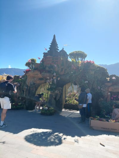 The image shows the entrance to the Ulun Danu Beratan Temple in Bedugul, Bali, Indonesia. The entrance is a large, ornate structure decorated with flowers and greenery, designed to resemble a stone archway. The temple's name is visible in text on the structure. Several people are present, with some posing for photos and others engaged in activities. One person, wearing a black t-shirt and white shorts, is taking a photo with their phone. Another person in a black top and skirt is seen bending down, possibly tending to plants. Two people in the background are facing the entrance, and another person is seated on the right side of the frame. The environment is outdoors, with a clear blue sky and mountains visible in the distance. The ground is paved with light-colored stone tiles, and shadows from trees are cast on the ground. There are also planters filled with flowers, and a cardboard box with text on it.