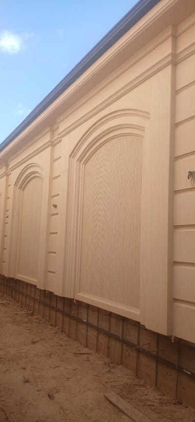 An outdoor, low-angle shot shows a cream-colored decorative wall under a bright blue sky with scattered white clouds. The wall features arched panels with vertical grooved detailing and horizontal bands. A dark-colored gutter runs along the top edge of the wall. The base of the wall is adjacent to a construction area with exposed earth and wooden supports. The image appears to be taken in Daliguzar, Uzbekistan.