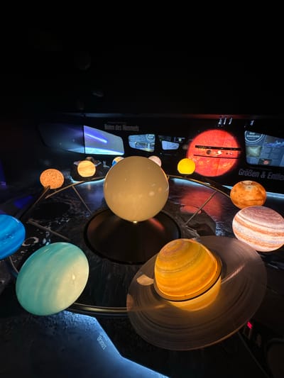The image depicts a display of the solar system, likely an educational exhibit. Several illuminated models of planets are arranged on a dark, circular base, suggesting a representation of planetary orbits. In the background, illuminated screens display astronomical imagery and text, with one screen showing a comet streaking across a starry sky, labeled "Boten des Himmels." Another screen, behind a bright red model resembling the sun, shows a diagram of the solar system with planets labeled, and text indicating "Unser Sonnensystem" (Our Solar System) and "Größen & Entfernungen" (Sizes & Distances).

One prominent model is a large, pale yellow sphere, possibly representing Earth or another terrestrial planet. Adjacent to it, a mottled orange sphere likely represents Mars. A large, striped sphere with distinct bands of color is a model of Jupiter, and another ringed sphere, glowing with warm yellow and orange bands, represents Saturn. Smaller, illuminated spheres in orange and yellow tones are also visible, likely representing inner planets or moons. A turquoise sphere, possibly Uranus, is in the foreground. Text projected onto the surface in front of the turquoise sphere reads "Uranus."

The exhibit appears to be located in a dimly lit indoor space, with the exhibit itself being the primary source of light. The overall ambiance is one of scientific or educational presentation. Without further context, the specific location in Salzburg, Austria, cannot be determined from the image alone.