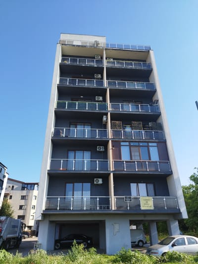This image captures a multi-story apartment building with a modern, dark-gray facade and glass balconies under a clear blue sky. The building features multiple levels, each with identical balconies. Air conditioning units are visible on several balconies. The ground level appears to be an open-air parking structure, with a black car and a white van parked underneath. Adjacent to the building, a white garbage truck with "URBAN" printed on its side is parked to the left. In the background, another building with a lighter facade and several windows is visible. A yellow sign with text reading "CABINET DE AVOCAT 0774 047 287" is affixed to one of the balconies. The scene is set in Bucharest, Romania, with lush green vegetation in the foreground.