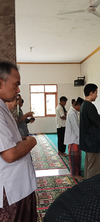 The image shows a group of men praying on prayer rugs in a mosque in Kecamatan Kutawaringin, Indonesia. The men are wearing traditional clothing and are standing in a row facing the direction of prayer. The walls are white and there is a window with a view of the outdoors. The ceiling is white and there is a fan hanging from the ceiling. The floor is tiled and covered with prayer rugs. The mosque is a simple, but well-maintained building. There is a speaker attached to the wall.  A man in a light blue shirt is standing in the foreground, his hands are crossed in front of him. He is looking to his left.  There is a man in a gray shirt in the background. He is looking towards the camera and he is holding his hands together in front of him.  The scene is peaceful and serene.