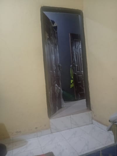 The image depicts an interior scene looking through an open doorway into another room. The primary focus is on a dark wooden door with ornate paneling, partially ajar. The doorframe and wall are a neutral, light yellow color. The floor in the foreground is tiled with a white marble pattern. Beyond the doorway, a second dark wooden door is visible, also slightly open. Inside the second room, a bright green object, possibly a vest or a piece of clothing, is hanging on the door. The lighting suggests an indoor setting, likely during the day, but the overall ambiance is somewhat dim. There is no visible text in the image. The scene appears to be a residential or interior space within Ekosa, Nigeria.