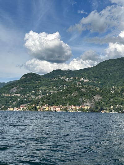 The image displays a scenic landscape depicting a body of water, a town, and mountainous terrain under a partly cloudy sky, characteristic of Gittana, Italy. The upper portion features a bright blue sky partially obscured by large, billowy white cumulus clouds, with some darker, shadowed clouds present towards the upper right. The middle ground is dominated by lush green mountains densely covered in trees and vegetation. Numerous residential structures, characterized by various colors and terracotta roofs, are scattered across the mountainsides, ascending from the shoreline. A distinct town or village extends along the base of the mountains at the water's edge, featuring closely packed buildings with colorful facades (yellows, oranges, and pastels) and a prominent, slender church tower or campanile on the left side of the town. The lower portion of the image shows the surface of a deep blue lake with visible ripples and small waves, suggesting slight movement or wind on the water. No individuals, specific actions, or interactions are discernible.