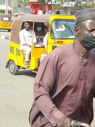 The scene depicts a daylight street in Babban Giji, Nigeria. In the immediate foreground, a dark-skinned male individual is partially visible, from the chest up, facing right. He wears a maroon kaftan with a patterned neckline, a dark kufi cap, and a black face mask covering his nose and mouth. His right hand grips a handlebar-like object, suggesting he is on a bicycle or motorcycle. Directly behind him, a yellow three-wheeled auto-rickshaw (keke Napep) with a brown fabric canopy travels right. The vehicle displays "KHC 506" in white text on its left side. Inside the keke Napep, a male passenger, likely the driver, wears a white kaftan and dark sandals, seated in the front and looking forward. Behind him, a second individual in dark attire wears a light blue face mask. A third, partially obscured individual is also visible within the vehicle. Further down the road in the background, another yellow keke Napep is visible. The street features asphalt pavement, a grey curb, and white line markings. Structures on the far side of the road include a building with a red facade and a "SHOW DECOR" sign. A red traffic light signal and green foliage are also present.
