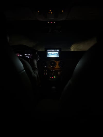 The image depicts the interior of a car at night. The dashboard and steering wheel are visible. A person is behind the wheel. A screen displaying an image is mounted on the dashboard. The location is unknown.