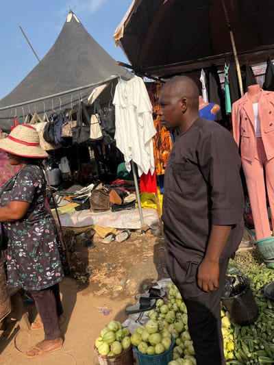 The image depicts a street market scene in Nyanya, Nigeria. Two individuals are visible. In the foreground, a person wearing a dark floral-patterned dress and brown trousers is sitting with their legs extended. They are also wearing a straw hat with red trim. Behind them, and to their left, a person wearing a dark brown tunic and black trousers is leaning forward, appearing to be looking at something below.
The scene is cluttered with various objects. In the background, racks of clothing, including pink jackets and pants, are displayed. Bags and other merchandise are also visible, some hanging from structures and others piled on the ground. Baskets filled with produce, likely okra and green, bulbous vegetables, are prominent. The ground is a mix of dirt and scattered debris. A black canopy tent provides shade.
The activity appears to be the daily operation of a market. The time of day seems to be during daylight hours, as indicated by the strong sunlight casting shadows. The weather appears to be warm and sunny. The overall impression is one of a busy, informal marketplace. No visible text is discernible in the image.