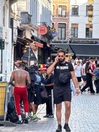 A man with a shaved head and a beard, wearing sunglasses and a black t-shirt with "BOXING" printed on it, is speaking on his phone while walking down a cobblestone street. He is also wearing a black fanny pack across his chest and black shorts. Behind him, a tattooed man in red track pants stands near a yellow barrier and several backpacks. In the background, several other people are walking, and buildings with shops and signs line the street. One sign reads "CITY MOBILO" and another has a red circle with a burger graphic. The atmosphere appears to be that of a busy city street, possibly in a commercial or tourist area. The architecture suggests a European city, and given the context, it is likely Brussels, Belgium, possibly in the Sainte-Catherine area due to the presence of a burger sign and what appears to be a food market setting.