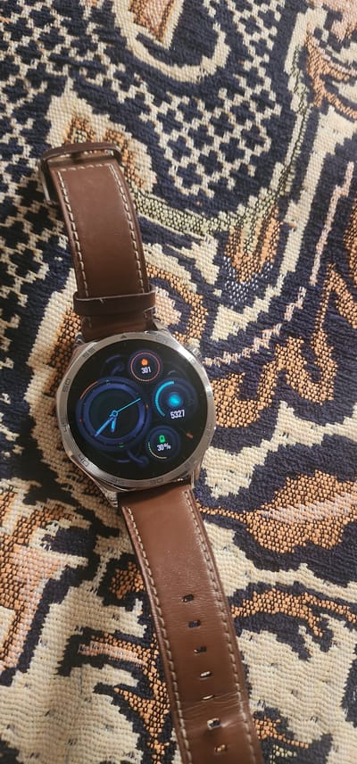 The image shows a close-up of a smartwatch with a brown leather strap lying on a patterned textile. The watch face displays several metrics: a circular graph with a pointer indicating 301, a digital readout of 5327, and a battery icon showing 30%. The textile has a complex geometric pattern in shades of dark blue, beige, and orange. The scene is lit from above, casting soft shadows. There is no indication of location within the city of Türkmenbaşy, Turkmenistan, as the image only depicts the smartwatch and the textile.
