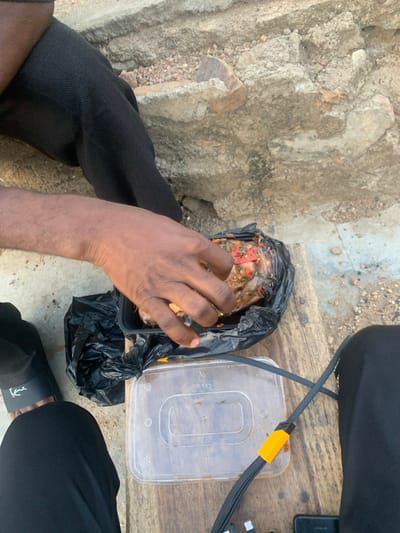 An overhead view captures a scene in Okene, Nigeria, featuring two individuals seated in an outdoor environment. The foreground consists of a weathered wooden surface, possibly a bench or table. In the immediate background is a rough, uneven concrete or rocky wall with exposed aggregate and dirt.
One individual's right arm and hand are prominent, reaching into a black rectangular plastic food container. Inside the container, a protein item, likely fish or meat, is visible, covered in a vibrant reddish-orange and green sauce containing visible spices or herbs. The food is partially encased within a crumpled black plastic bag lining the container. This individual wears black trousers and a dark-colored slide sandal with "Kane" visible on the strap.
To the upper left, a portion of another person's leg, also clad in black trousers, is visible.
On the wooden surface, positioned below the active food container, is a clear rectangular plastic container with a lid, appearing mostly empty. Adjacent to it, a black braided charging cable with a distinctive yellow plastic segment is laid out, connecting to or resting near a dark-colored smartphone or small electronic device partially visible on the right edge of the frame.