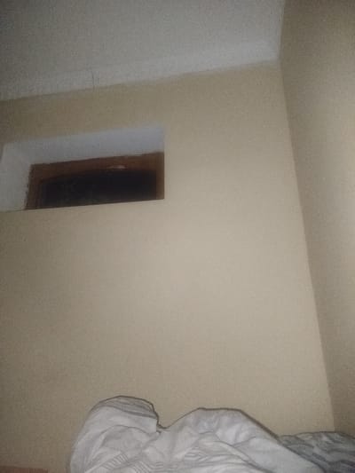 The image depicts a corner of a room. A wall is painted a light tan color. There's a rectangular recess in the wall, possibly a window or niche, with a darker interior. The top of the recess is painted white. In the lower portion of the image, there is a bed covered with a white and gray patterned blanket. The ceiling is white and has a small crack where the wall meets it. There's nothing to indicate the location is in Chumbagish, Uzbekistan; that information is not discernible from the image.