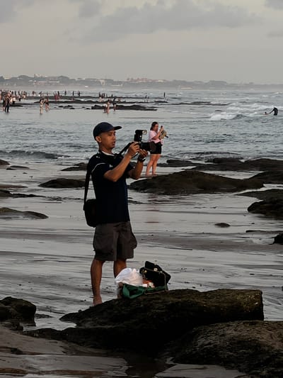 A man is taking photographs on the beach at Canggu, Indonesia. The man is standing barefoot, wearing a dark blue shirt, gray shorts, and a baseball cap. He holds a camera to his eye. In the background are other people at the shore, and rock formations along the beach. Further in the background are buildings along the beach on the shore. The sky is overcast.