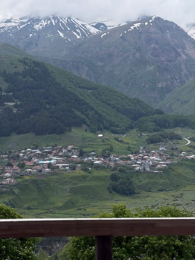 This media file depicts a high-angle view of a village nestled in a mountainous landscape. The scene is dominated by rolling green hills and dramatic, rocky mountains in the background, some of which are dusted with snow. A dense cluster of houses with red and white roofs forms the village, spread out along the contours of the terrain. A winding road is visible, snaking through the village and the surrounding landscape. The overall setting suggests a rural, mountainous area, likely in a temperate climate given the lush greenery.
The time of day appears to be daytime, with soft lighting suggesting either early morning or late afternoon. The weather seems overcast or hazy, as indicated by the diffused light and the presence of clouds or mist around the snow-capped peaks. There are no visible people or explicit activities taking place, creating a sense of stillness and tranquility. The presence of trees and vegetation suggests a natural, possibly remote location. The image also includes a wooden structure in the foreground, possibly a balcony or fence, which frames the view and adds a sense of perspective. No text is visible in the media file.