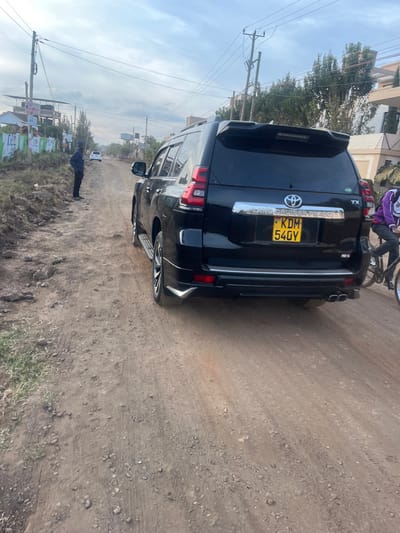 The image, captured in what appears to be portrait orientation and presented rotated, shows the rear and side of a black Toyota Land Cruiser Prado SUV. The vehicle is positioned on a dusty, unpaved road that dominates the right side of the frame and extends towards the background.
**Subjects and Objects:** The black SUV is a central subject, displaying a Kenyan license plate "KDM 540Y" on its tailgate. The model badges "LAND CRUISER PRADO" and "TX" are clearly visible. The vehicle features dual exhaust pipes on its right rear side. Near the SUV's rear, a person wearing a purple jacket is seen riding a bicycle. Further down the road, to the bottom-left of the SUV's current position, another person in dark attire is standing beside a series of posters or banners lining the roadside. Utility poles with multiple wires are visible along the left side of the road, with buildings and sparse trees in the background.
**Scene and Setting:** The setting appears to be a semi-urban or rural area, characterized by the unpaved, dry, and dusty road surface. The sky is clear and bright, indicating daytime and sunny, dry weather conditions.
**Activity:** The SUV appears to be either stationary or moving slowly. The person on the bicycle is actively riding, while the other person is standing still.
**Notable Details:** The "KDM 540Y" license plate confirms the location is Kenya, aligning with the provided context of Gitothua ward. The general appearance of the infrastructure and environment also suggests an East African setting. No specific emotions are discernible from the subjects.