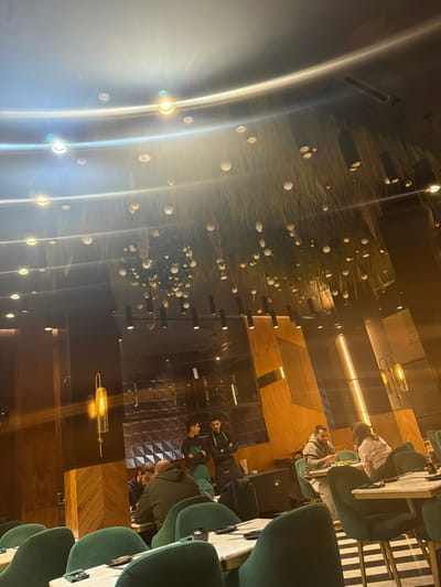 Indoor scene features diners seated in plush green chairs at small tables. Behind them, a dark wall has decorative elements including a tree-like structure with white spheres and a series of suspended lights. Light fixtures illuminate the area, creating reflections on the glossy surfaces. Some individuals are partially visible, suggesting an occupied space. The restaurant has modern design and a relaxed atmosphere.