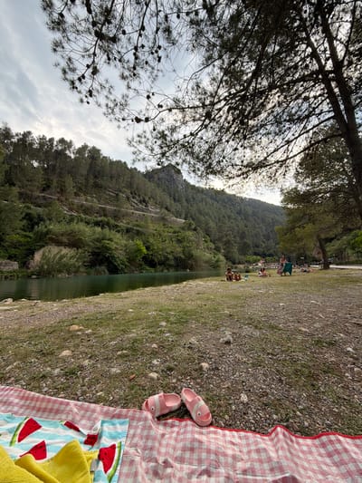 The scene is set on the bank of a river at Balneario Fuente de Baños Montanejos, a thermal spring area in Montanejos, Spain. The view looks toward a steep, tree-covered mountain. A picnic blanket with a red and white checkered pattern is visible in the foreground, with a pair of pink sandals and a blue towel with watermelon prints placed on it. A yellow towel is also partly visible. Several people are on the opposite bank of the river, some sitting, some standing, and some near the water's edge. A dense canopy of trees stretches across the top of the frame.