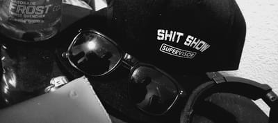 The image shows a black and white photo of various objects: a black cap with the words "SHIT SHOW SUPERVISOR" stitched on, a pair of sunglasses, a half-empty bottle of Gatorade Frost, an object resembling a lighter, and headphones, likely on a table or a similar surface. The objects are positioned in a cluttered manner. The coordinates 37.776, -122.421 correspond to a location in San Francisco, United States. The precise location within San Francisco is not available in the image.
