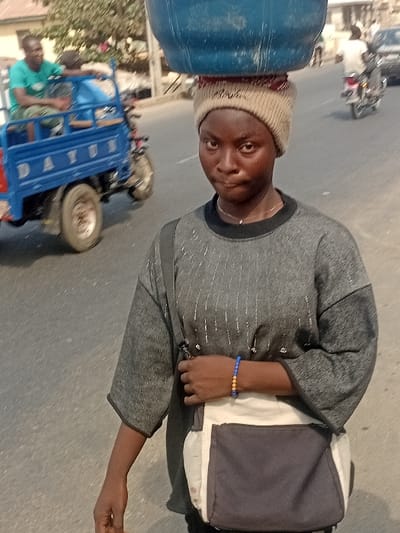 A person carries a large blue water jug on their head, wearing a beige knit hat and a gray, long-sleeved shirt. A messenger bag hangs from one shoulder. The person stands on what appears to be a road. In the background, a blue three-wheeled vehicle with two men in it is slightly blurred, along with other vehicles and indistinct buildings. The scene seems to be set in a bustling, possibly urban environment.