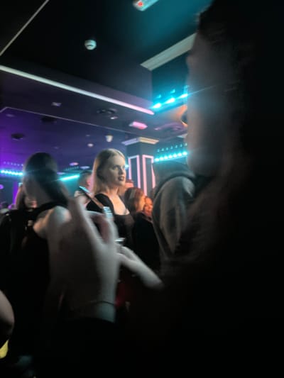 The image depicts an indoor scene, likely a nightclub or similar venue, characterized by low lighting and colorful illumination from neon lights and stage lighting. In the foreground, a person's hand holding a drink is partially visible, suggesting an active social setting. Further back, several individuals are present, with one woman in a black top standing out due to her lighter complexion and blonde hair. Another person wearing a grey hoodie is also visible. The environment features a dark ceiling with what appear to be smoke detectors and recessed lighting. The overall impression is a candid shot taken during a social gathering in a dimly lit, entertainment-focused location. There is no information to determine the location in the city of Kirov, Russia.
