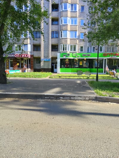 The image captures a street view in Abakan, Russia, on a sunny day, featuring the facade of a multi-story brick apartment building with ground-level commercial establishments. A tree with lush green foliage partially obstructs the view on the left. The ground level of the building houses two visible shops: one on the left with a red and brown facade, displaying signage for "UNIVERSAL OPTIKA" and "ОПТИКА," and an entrance with a ramp and railings. Another shop to the right, with a bright green facade, is identified as "ОПТИК ПЛЮС" and features large display windows showcasing mannequins with eyewear and promotional posters. A blue sign for "СРОЧНЫЙ РЕМОНТ ОБУВИ" (Urgent Shoe Repair) is visible between the two shops. In front of the "ОПТИК ПЛЮС" store, a black metal fence separates the sidewalk from a strip of green grass. Two large, circular metal sculptures resembling eyeglasses are placed on the grassy area. A black lamppost stands near the fence. Further to the right, a person is seated on a park bench, facing away from the camera, looking towards the building. The foreground is dominated by a paved road with a raised concrete curb separating it from the sidewalk and grassy area. Shadows from the tree and the building are cast across the road and sidewalk, indicating the time of day.