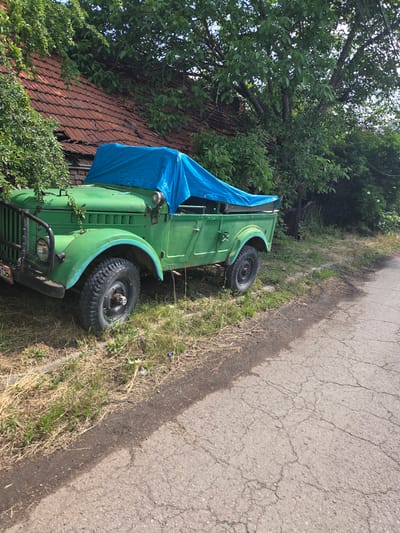 The image displays an outdoor scene in Elin Pelin, Bulgaria, on a clear or partly cloudy day. The primary subject is a faded green, possibly military-style, utility vehicle parked on an overgrown grass and dirt verge beside a cracked asphalt road. No individuals are visible.

The vehicle, appearing to be an older model 4x4, features a robust design. Its front left section is prominently visible, showing a round headlight, a black tubular grille guard, and a fragment of a white license plate with black characters, "OCB" being legible. The vehicle's paint is a worn green, with hints of a lighter blue paint present on the front left fender above the wheel. The front left wheel is equipped with a chunky, off-road tire. The open-top rear section and parts of the cabin are covered by a wrinkled, bright blue tarpaulin. Green plant tendrils are seen growing over the vehicle's front fender.

Behind the vehicle, an old building with a reddish-brown tiled roof is partially obscured by dense green foliage. The terracotta roof tiles exhibit wear and damage, with some appearing broken or missing. Large, mature deciduous trees with lush green leaves dominate the background and upper sections of the image, contributing to the overgrown appearance of the area surrounding the building.

The ground where the vehicle is parked consists of dry, yellowish grass mixed with green weeds and exposed soil. A distinct, uneven edge separates this unkempt verge from the adjacent road. The road surface is asphalt, severely weathered with a network of cracks and fissures across its visible expanse. The lighting suggests direct sunlight, creating subtle shadows.
