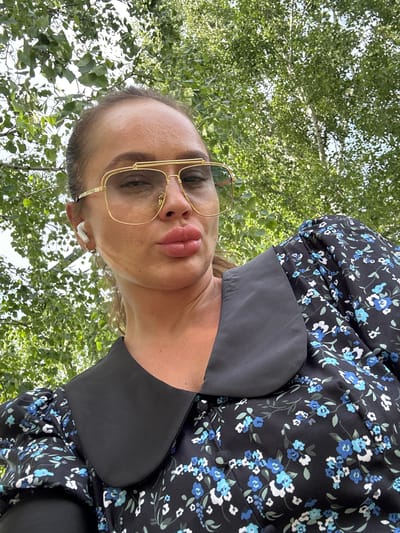 The image is a close-up, slightly tilted selfie of a woman in an outdoor setting. The woman is wearing large, gold-rimmed glasses and has full, pink lips. She is also wearing white wireless earbuds. Her dress is black with a floral pattern featuring blue and white flowers. A dark fabric collar or neckline is visible on the dress.

The background consists primarily of lush green foliage, suggesting a natural, possibly wooded, environment. The lighting indicates it is daytime, with sunlight filtering through the leaves. There are no visible text elements in the image. The woman's expression is neutral. The context provided places the image in Yuzhnouralsk, Russia.