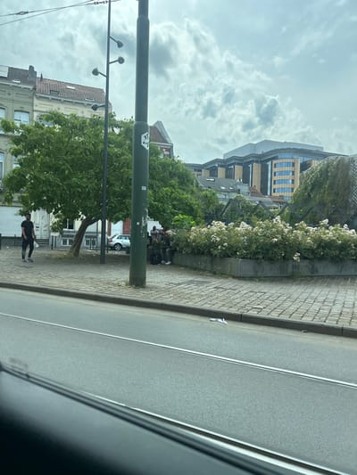 The image, taken in Anderlecht, Belgium, shows a street scene. In the foreground, a tram track runs parallel to the road. On the sidewalk, a young man in a black t-shirt and black pants stands holding a white plastic bag. Further down the sidewalk, a group of people are seated on a planter, some with backpacks. Behind them, a silver car is parked. The sidewalk is paved with cobblestones. Trees line the sidewalk, and behind them, buildings of various architectural styles are visible, including a modern glass-fronted building. The sky is overcast with clouds.