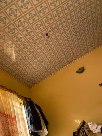 The image depicts the interior of a room in Kanshio, Nigeria. The ceiling is covered with decorative tiles featuring a repeating geometric pattern of light gray, light blue, and beige squares and diamonds. A single dark-colored hook is affixed to the ceiling, slightly off-center. The walls are painted a solid yellowish-orange color. On the right wall, a semi-circular wall-mounted light fixture with a translucent green and white cover is visible, along with minor cracks in the paint. Towards the bottom right, a pile of folded clothing, including a light brown garment and a white garment, rests on an unseen surface. On the left side, a window is partially visible, covered by light-colored curtains with a vertical stripe pattern. A dark-colored jacket or blazer hangs on a hanger from a rod near the window, with other light-colored garments visible beneath it. No individuals or active interactions are present in the scene.