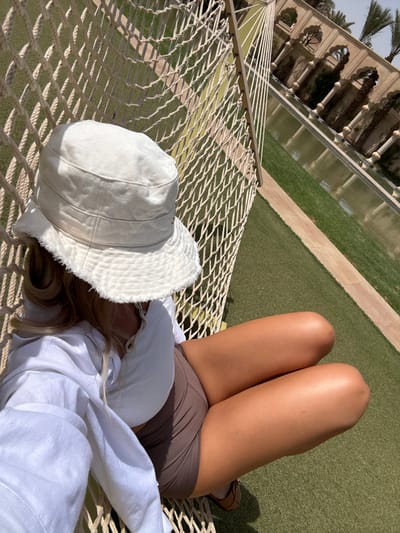 A young woman wearing a white shirt, brown shorts, and a white bucket hat sits in a hammock. The hammock is strung between two posts. The woman is looking down at her phone. The scene is located in Ouled Jelal, Morocco at coordinates 31.664, -7.871. The backdrop features an ornate, arched structure that surrounds a rectangular pool of water. The pool is surrounded by manicured green grass, and the structure is made of white stone and has a traditional Moroccan architectural design. The scene is bathed in sunlight.