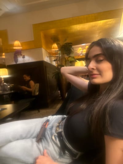 A young woman with long, dark hair is sitting at a table, leaning on her left arm. She is wearing a dark top and light blue jeans with a belt. A mobile phone is partially visible in her front pocket. The background features a reflective surface, likely a table or counter, showing a blurred reflection of another person and decorative elements, including lamps and plants. The location is likely a hotel or lounge area in the city of Bellevue, Switzerland, suggested by the interior design and the presence of decorative lamps and upscale furnishings.