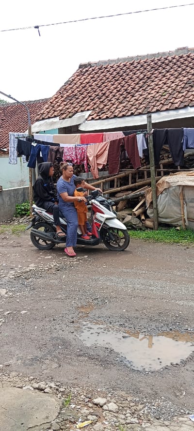 The image depicts a scene in Kutawaringin, Indonesia, with coordinates -6.974, 107.519. Three individuals, including a woman and two children, are situated on a white Yamaha motorcycle. The woman is wearing a brown shirt and blue pants, while the children are wearing orange shirts. The motorcycle is parked on a dirt road in front of a residential building with a red tiled roof. Several pieces of laundry are hanging on a clothesline in front of the building. The road is rough and uneven, with a small puddle of water reflecting the surrounding environment. The scene suggests a typical residential area in a developing country.