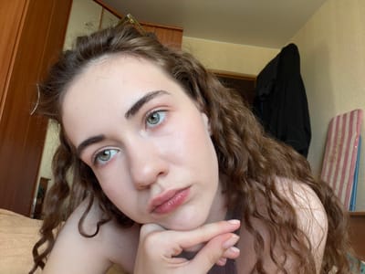 The image shows a close-up of a young woman with long, curly brown hair and green eyes. She is resting her chin on her hand and looking away from the camera with a pensive expression. The background suggests an indoor setting, possibly a bedroom, with a wooden wardrobe on the left and a wall with a coat hanging on it on the right. A striped object, possibly a board or fabric, is visible against the wall. The lighting appears to be natural, coming from a window or overhead source. There are no specific landmarks or indicators that place the scene in Zavodoukovsk, Russia.