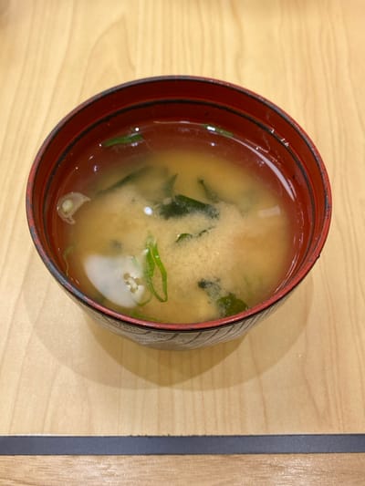 A top-down shot features a bowl of miso soup resting on a light-colored wooden table. The bowl is dark red with a black exterior, showing some signs of wear or texture along its rim. Inside, the soup is a cloudy, yellowish-brown broth, characteristic of miso. Visible ingredients include several pieces of dark green wakame seaweed, thin slices of bright green scallions or green onions, and irregular white pieces of tofu, some of which exhibit small textural holes.

The wooden table surface has a fine grain pattern and a matte finish. To the right of the bowl, a distinct dark grey, almost black, strip runs vertically across the table, suggesting it might be part of a placemat, a table runner, or a division in the table's surface. A soft shadow is cast by the bowl onto the table, indicating a light source from the upper left.

The scene is static, depicting food ready for consumption. There are no people visible, and no activity is taking place. The indoor setting provides no specific cues for time of day or weather. There is no visible text in the image. The bowl is positioned slightly off-center and the image appears to be slightly rotated.