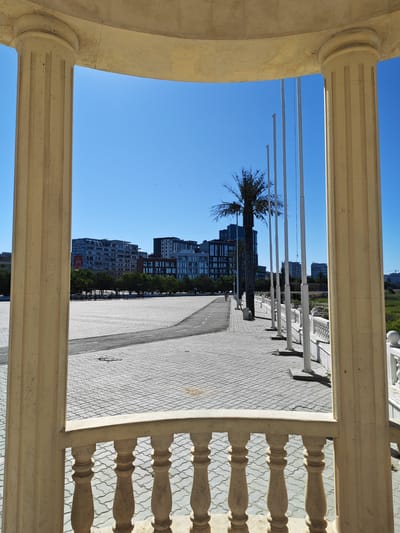 The image captures a sunlit scene framed by classical stone columns and balustrades, looking out onto an urban landscape in Aktau, Kazakhstan. In the foreground, the ornate stone balustrade and fluted columns suggest an architectural feature like a pavilion or belvedere. Beyond this, a wide, paved plaza stretches out, constructed with light-colored interlocking stone tiles. A paved road or pathway curves through the plaza.
In the mid-ground, a row of tall, thin flagpoles stands against the clear blue sky, some appearing to be without flags. Adjacent to the flagpoles is a solitary palm tree, its fronds silhouetted against the bright backdrop. Behind the palm tree and flagpoles, a line of trees forms a verdant barrier.
The background is dominated by modern, multi-story buildings of varying designs and colors, indicative of a city environment. Some buildings have a light grey facade with numerous windows, while others feature darker tones and more contemporary architectural styles. The sky is a uniform, vivid blue with a few distant, barely visible specks that could be birds or distant aircraft. The overall impression is one of a well-maintained public space in a coastal city.
