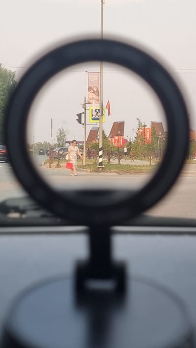 The image shows a view through a circular lens, possibly a camera lens or a magnifying glass, onto a street scene. The scene appears to be in an outdoor setting, likely a roadside area in a town or city. The perspective is from a vehicle, as indicated by the reflection of a car mirror on the right side of the image.
In the foreground, within the circular frame, a person is visible in a light-colored dress, carrying a red bag. They are standing on the side of a paved road. Behind them, there are traffic signs, including a pedestrian crossing sign. Further in the background, there are buildings with distinctive red triangular roofs and some trees. Several signs with text are visible. One banner on a pole has an image of what appears to be a person and some text. The buildings have large red signs with white text that seems to read "ПОБЕДА!". The sky is overcast, suggesting a possibly cloudy or hazy day. The overall impression is a casual daytime scene, possibly in a commercial or residential area. The orientation of the image is rotated, making the "upside down" perspective a notable detail.