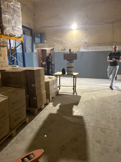 The image depicts an indoor storage area, possibly a warehouse, located in Dupnitsa, Bulgaria. The scene is cluttered with cardboard boxes stacked on wooden pallets, some of which are wrapped in plastic. In the foreground, an orange pallet jack is visible. In the mid-ground, a man wearing a dark t-shirt and grey sweatpants stands near a table with stacked buckets and what appears to be a wooden structure on top. Another man, wearing a dark t-shirt and dark pants, is seen in the background near a doorway. The environment appears utilitarian, with bare walls and exposed electrical conduits. Lighting is provided by an overhead bulb.