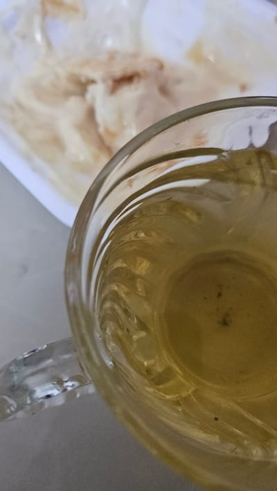 A top-down shot depicts a glass cup filled with a yellowish liquid, possibly tea, placed on a white surface. To the left, a blurry, white plate holds food with a mottled tan and white appearance. The glass cup has a clear, ribbed design. The scene is in Cam Đức, Vietnam.