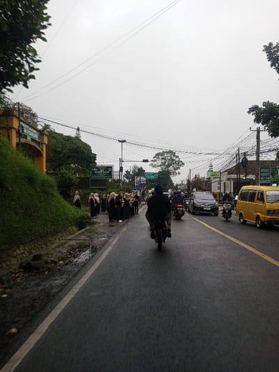 The image portrays a wet, overcast street scene in Chagzam_Pam, Bhutan. The asphalt road, marked by a solid white line on the left shoulder and a solid yellow center line, shows visible moisture from recent rain. A person on a black motorcycle, wearing a dark jacket, is in the foreground, traveling away from the camera. Multiple other motorcycles and vehicles, including a dark SUV and a yellow van, are visible in both directions, forming traffic.
On the left side of the road, a group of approximately 10-15 individuals, predominantly women, stand on the concrete shoulder. They are dressed in black garments and various light-colored headscarves (hijabs). Behind them, a steep, green grassy embankment rises, partially obscuring a yellow archway structure and a prominent green sign displaying Indonesian text, "SMA MANBAUL 'ULUM" and "MENERIMA SISWA BARU." A small stream of water flows in a gutter at the base of the embankment.
Utility poles support a dense network of overhead electrical wires across the frame. Buildings line the right side of the street, with a prominent yellow sign featuring black text "AYAM PENYET KANTIN" visible on one structure. The sky is uniformly light grey, indicating heavy cloud cover. Traffic lights are visible further down the road.
