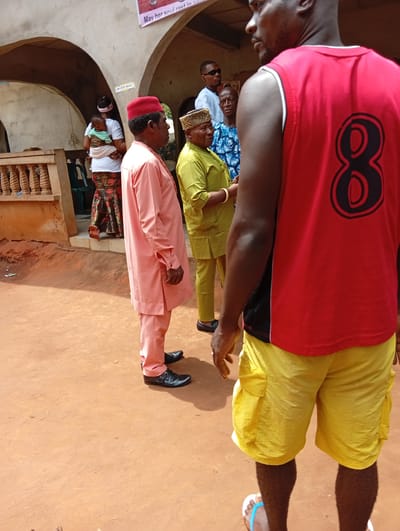 The image captures a scene in Umundugba, Nigeria. In the foreground, a man in a red basketball jersey with the number 8 and yellow shorts stands with his back to the camera. A man in a peach-colored traditional outfit and red cap stands to the left, facing the group. To the right of the man in peach is another man in a green traditional outfit and a decorated hat.
Further back, additional individuals are visible, including a man in sunglasses and a blue patterned shirt. A woman is carrying a baby.
The setting appears to be outdoors, possibly near a building with arches. The ground is reddish-brown earth. A sign is visible above the group of people, with the words "May her soul rest in".