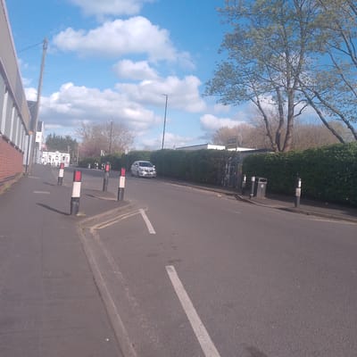The image depicts a street in Coventry, United Kingdom. The scene includes a road with white dashed lines, a sidewalk, and a building on the left side. There are bollards with alternating red/black and white/black stripes along the edge of the sidewalk. A white car is visible on the right side of the road, adjacent to a green hedge. Behind the hedge, there is a sign that says "Southfields Primary School, Main Reception". The sky is blue with scattered white clouds. Trees are visible in the background.