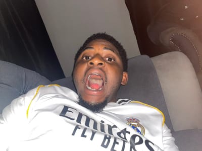 The image depicts an adult male situated indoors, stated to be in the city of Ube, Nigeria. The subject, with dark skin, short dark hair, and a goatee beard, is positioned centrally and looking directly at the camera. His mouth is open wide, revealing his tongue and the oral cavity. He is wearing a white short-sleeved shirt featuring the 'Emirates FLY BETR' sponsor logo and the Real Madrid club crest, with yellow detailing on the collar or shoulder seam. The individual is seated on a dark grey upholstered chair or sofa. In the background to the right, a section of another dark upholstered furniture piece, adorned with studded detailing, is partially visible. To the left background, a dark, indistinct surface, possibly a curtain or wall, is present. The scene is illuminated, highlighting the subject's face against a generally dim background.