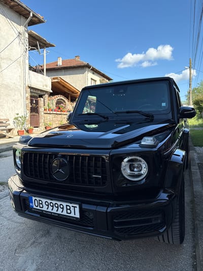A black Mercedes-Benz G-Class AMG SUV is parked on a concrete street in Lukovit, Bulgaria. The car's license plate reads "OB 9999 BT". The front of the vehicle faces the camera, showcasing its grille, headlamp, and Bulgarian license plate. In the background, buildings with tiled roofs and stone facades are visible, along with a clear blue sky with a few clouds and overhead power lines. The buildings suggest an older style of architecture. The SUV is parked near the edge of the street.