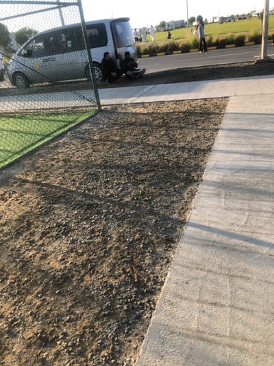 The image captures an outdoor scene in the city of النعيلة, Saudi Arabia, during daylight hours, indicated by strong directional lighting and long shadows. The immediate foreground on the left contains a green chain-link fence, behind which artificial green turf is visible. Adjacent to the fence, a patch of bare ground composed of dirt and scattered gravel extends towards a clean concrete pathway that occupies the right side of the frame. In the midground, a silver minivan is parked parallel to a paved road. Two adult males are seated on the ground next to the driver's side of the minivan. The male on the left wears dark clothing. The male on the right, also in dark attire, holds an object in his hands and has a white rectangular object on the ground beside him. The paved road features a yellow and black striped curb, bordering a landscaped median with low green shrubs. The background reveals a wide, open green field where approximately ten individuals are present, some standing and others seated, suggesting recreational activity. Several individuals on the field are dressed in traditional white thobes. Additional vehicles are visible in the far background.